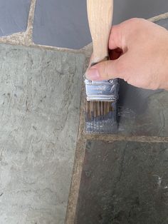 a hand holding a paintbrush over a gray tile floor with grey grouting