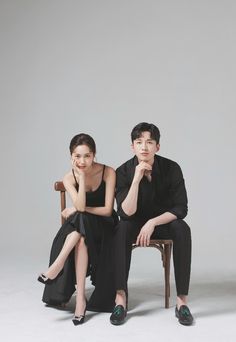 a man and woman sitting on top of a wooden chair next to each other in front of a gray background