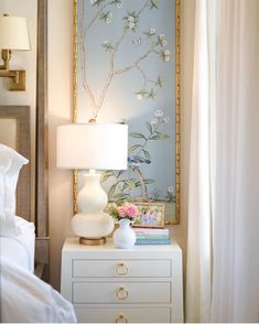 a white nightstand with flowers on it next to a painting and lamp in a bedroom