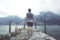 a man standing on a pier looking out at the water with mountains in the background