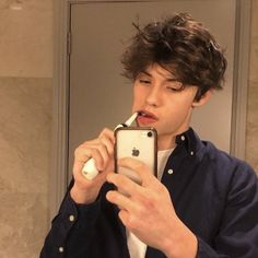 a young man brushing his teeth in front of a mirror while looking at his cell phone