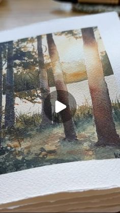 an open book with watercolors on it and a painting of trees in the background