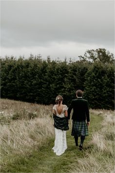 a bride and groom walking through tall grass in front of trees on a cloudy day