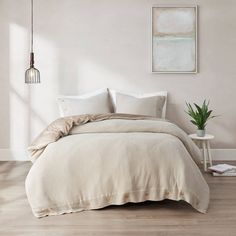 a bedroom with white walls and wood flooring has a large bed covered in beige linens