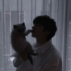 a man holding a cat in his right hand while standing next to a window with white curtains