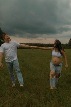 a pregnant woman holding the hand of a man in a field with dark clouds overhead