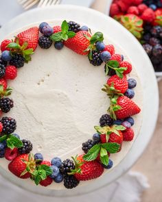 a cake decorated with berries and mint leaves on a white plate next to a bowl of strawberries