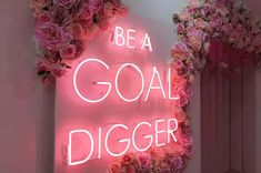 a pink neon sign that says be a goal digger surrounded by flowers and greenery