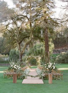 an outdoor ceremony set up with wooden chairs and floral arrangements on the grass, surrounded by trees