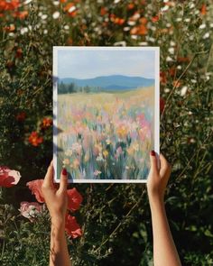 a person holding up a painting in front of flowers and grass with mountains in the background