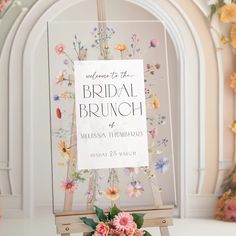 a sign with flowers on it sitting in front of a floral arch for a bridal brunch