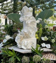 a white wedding cake surrounded by flowers and greenery in a garden center with palm trees