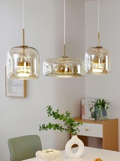 three glass pendant lights hanging over a dining room table with two chairs and a potted plant