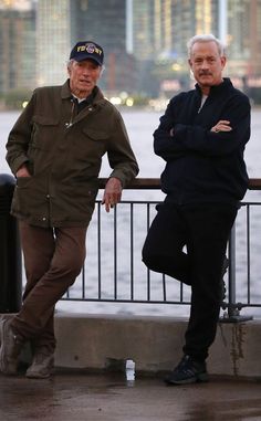 two older men standing next to each other on a bridge near the water with buildings in the background