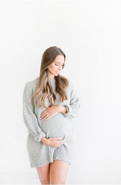 a pregnant woman wearing a grey sweater dress and holding her stomach in front of a white wall