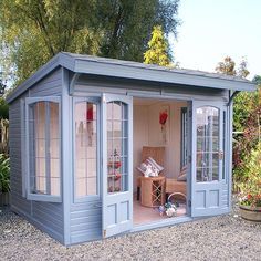 a small blue shed with windows and doors on gravel ground next to trees, shrubs and flowers