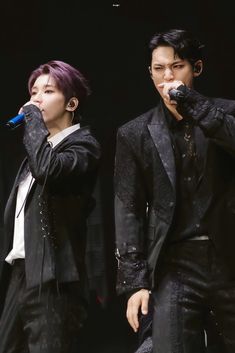 two people standing next to each other with microphones in their hands and wearing black suits