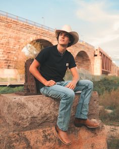 a young man wearing a cowboy hat sitting on a rock in front of a bridge