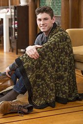 a young man sitting on the floor wrapped in a knitted blanket and smiling at the camera