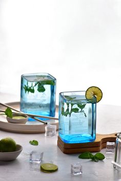 two glasses filled with water sitting on top of a table next to limes and spoons