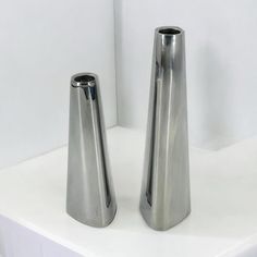 two silver vases sitting on top of a white table next to each other in front of a white wall