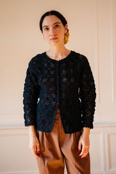 a woman standing in front of a white wall wearing brown pants and a black knitted jacket