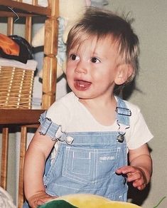 a toddler in overalls standing next to a plate with food on top of it