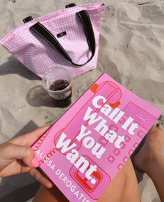 a person holding up a pink book on the beach with a tote bag next to it