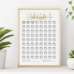 a white framed poster with gold foil numbers and houses on it next to potted plants