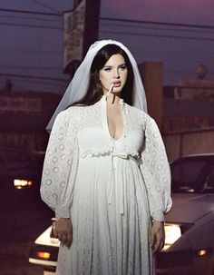 a woman in a white wedding dress standing next to a car at night with her hands on her hips