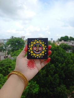 a person holding up a small square object in front of the camera with trees and buildings in the background