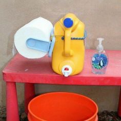 a red table topped with two orange buckets next to a yellow and white toilet paper dispenser