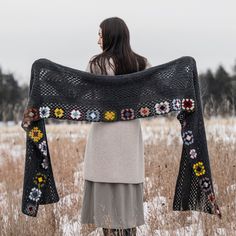 a woman standing in a field holding up a black crocheted shawl with flowers on it