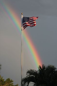 a rainbow shines in the sky over a flag pole and palm trees with a dark gray sky