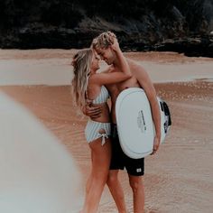 two people are hugging on the beach with a surfboard in their hands and one person is wearing a bathing suit