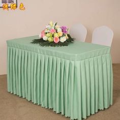 the table is covered with a green cloth and has flowers on it, along with two white chairs