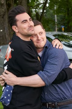 two men hugging each other in front of trees and parked cars on the side of the road