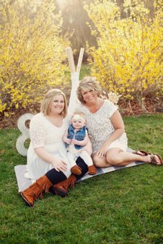 two women and a baby are sitting on the grass in front of some bushes with yellow flowers