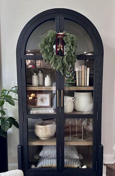 a black china cabinet with wreath on top and dishes in the glass front, sitting next to a potted plant