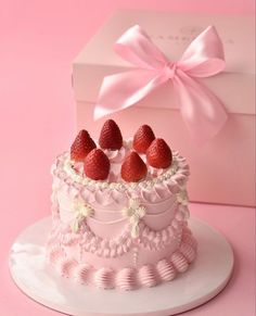 a pink cake with strawberries on top in front of a gift box and ribbon