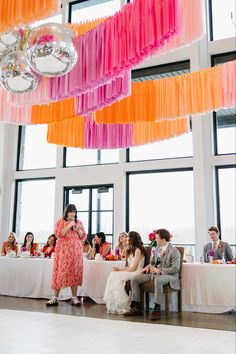 a woman standing in front of a group of people at a table with orange and pink streamers hanging from the ceiling