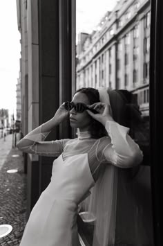 a black and white photo of a woman in a dress leaning against a wall with her hands on her head