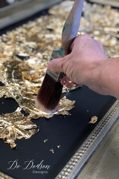 a person holding a paintbrush painting gold leaf designs on a black tray with silver trim