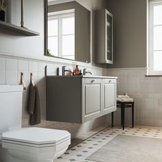 a white toilet sitting next to a bathroom sink under two windows in a room with tile flooring