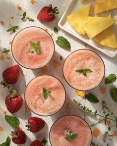 three glasses filled with smoothies next to sliced pineapple and strawberries