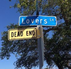 black and white photograph of two street signs that read lovers in dead end with trees in the background