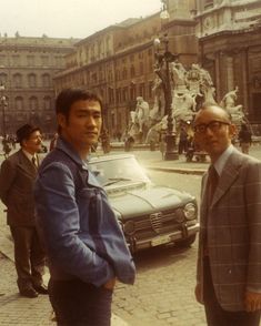 two men standing next to each other in front of a car on a city street