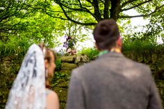 a bride and groom looking at each other in front of a stone wall with trees