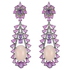 Step into a world of botanical elegance with our Victorian 27.85 Cttw. Peach Coral, Diamond, and Multi Sapphire Floral Dangle Earrings. A true floral masterpiece, these earrings capture the essence of a blooming garden with meticulous craftsmanship and a harmonious blend of precious gemstones. The surmount is a captivating floral arrangement, with a central core surrounded by a halo of sparkling white diamonds, reminiscent of a delicate flower in full bloom. The long drop, crafted in black rhodium and adorned with glistening diamonds, extends gracefully to cradle a cabochon peach coral. This coral, with its smooth, rounded surface, adds a touch of warmth and uniqueness to the design. The intricate floral setup is completed by pink pear-shaped sapphires connecting the diamond frames, creati Blooming Garden, Multi Sapphire, In Full Bloom, Black Rhodium, Delicate Flower, Floral Earrings, Floral Arrangement, Precious Gemstones, White Diamonds