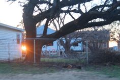 the sun is setting behind a chain link fence in front of a small white house